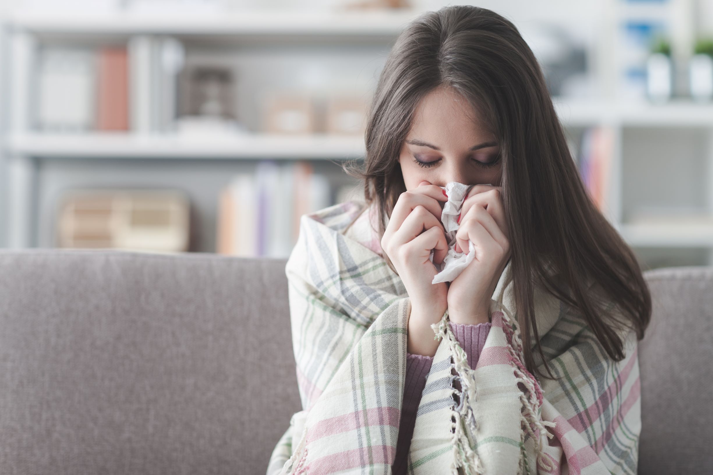 Woman suffering from cold and flu symptoms, putting paper tissue on her nose. Sitting on couch and covered with blankets.