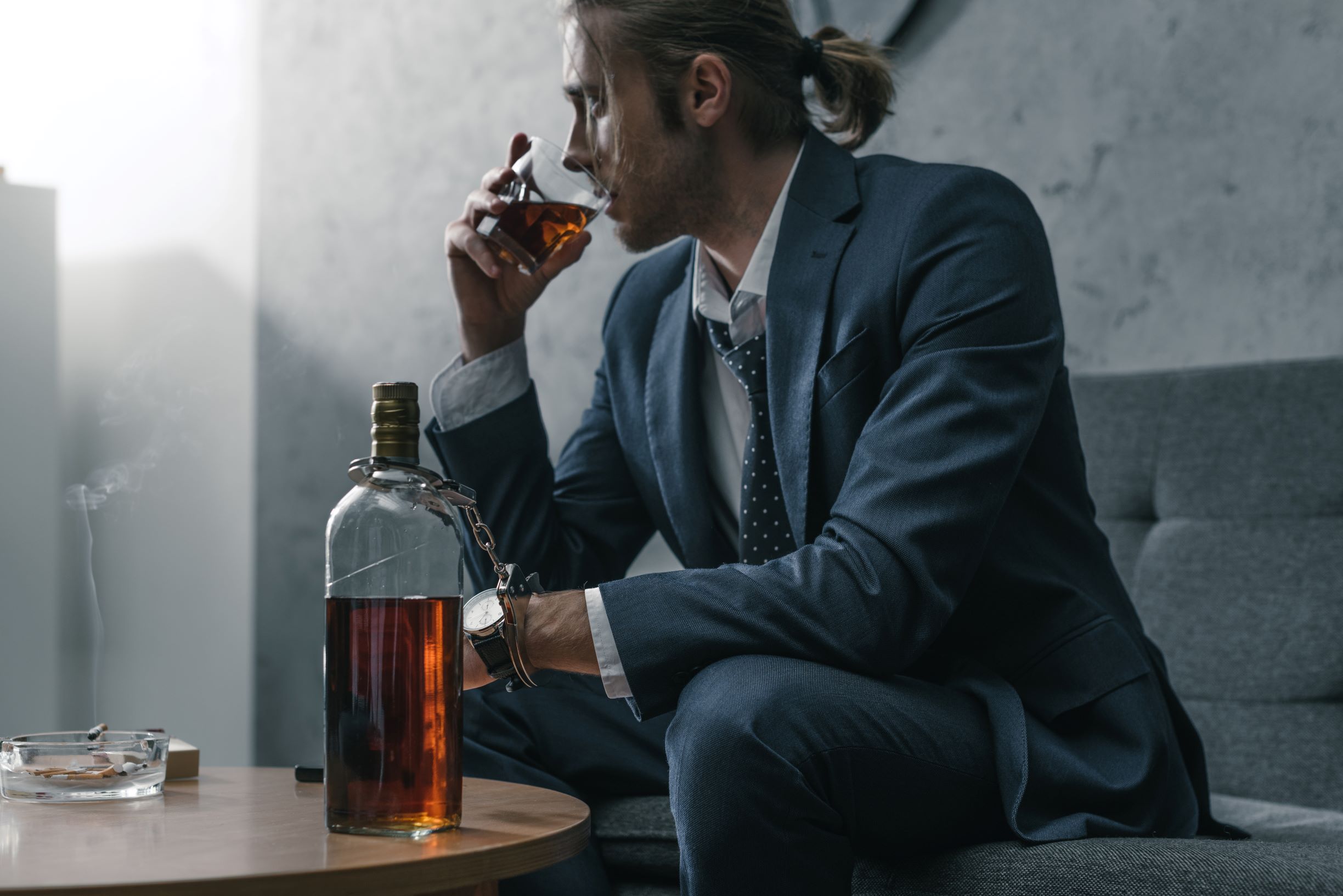 Man suffering from alcohol addiction, sitting and drinking alcohol from a glass. In front of him a big bottle of whiskey.