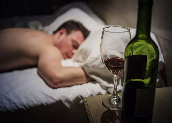 Man sleeping in bed on his face, suffering from hangover symptoms, with a bottle of wine and a glass next to him.