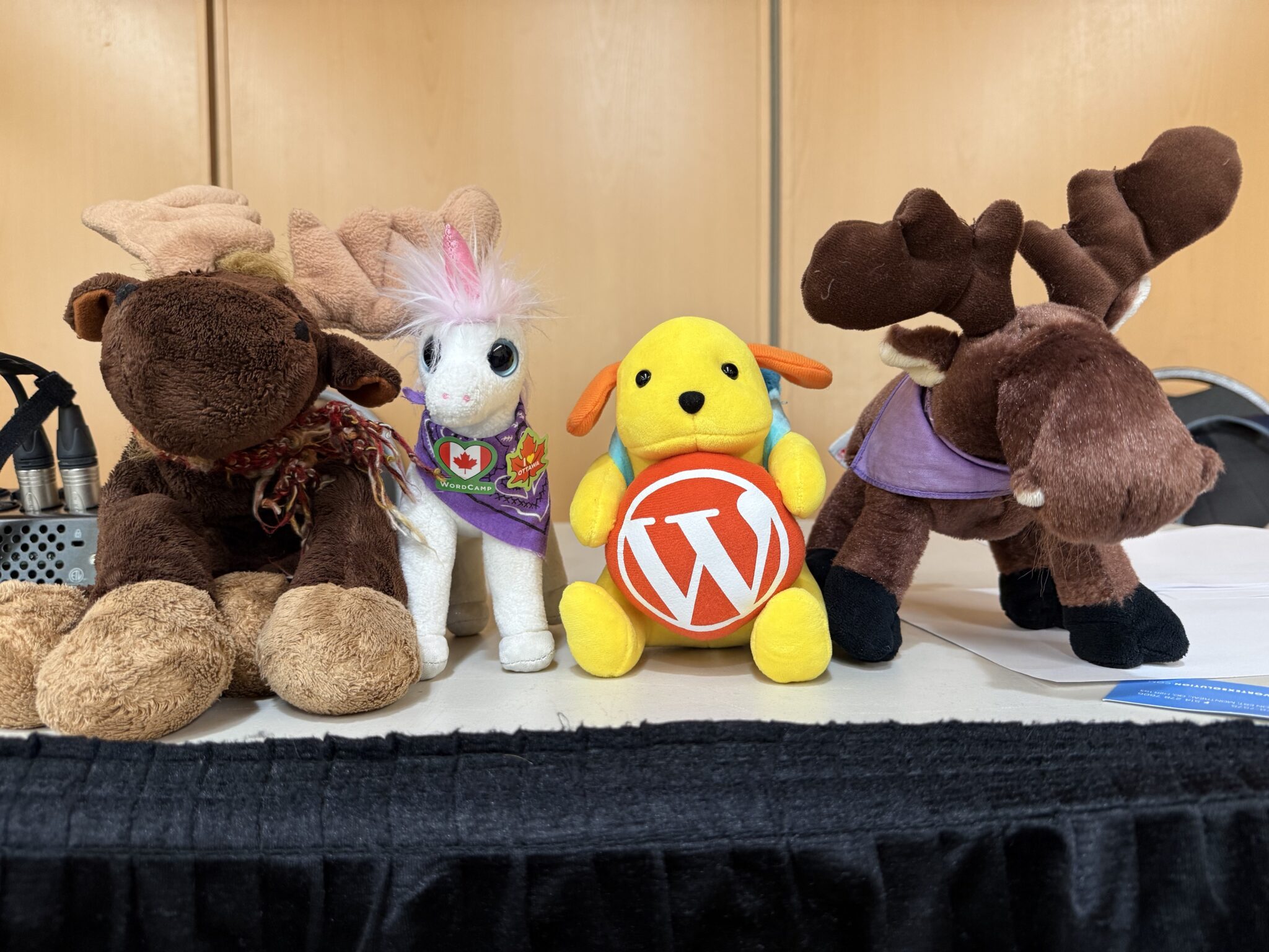From left to right, a stuffed moose, a stuffed unicorn with Canada charms (a Canadian flag inside a heart and a maple leaf), a Wapuu with an orange WordPress logo and another moose standing next to each other on a table at WordCamp Canada.