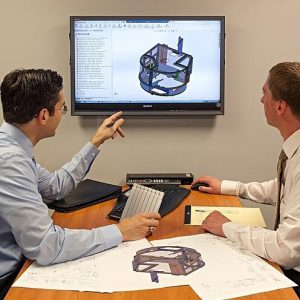 Program managers and engineers meeting to perform a production readiness review on a CAD model seen on the projector screen. 