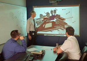 Program managers and engineers meeting to perform a production readiness review on a CAD model that's seen on the projector screen. 