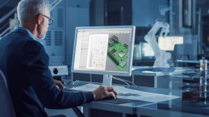 Engineer Works on Computer Uses CAD Software to Design 3D Industrial Machinery Component. In the Background Robot Arm Concept Standing in Heavy Industry Engineering Facility. Over the Shoulder Shot