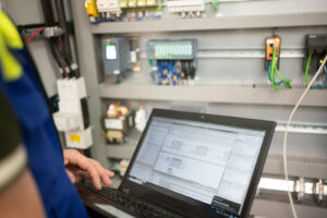 Programmer checking for correct function of PLC programming with electrical cabinet in background