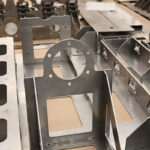 Close-up of laser-cut and formed steel brackets with circular and rectangular cutouts, staged for welding in a sheet metal fabrication shop.