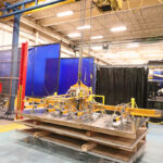 Amada sheet stacker with vacuum lifter on an overhead crane handling metal sheet pallets in a fabrication shop.