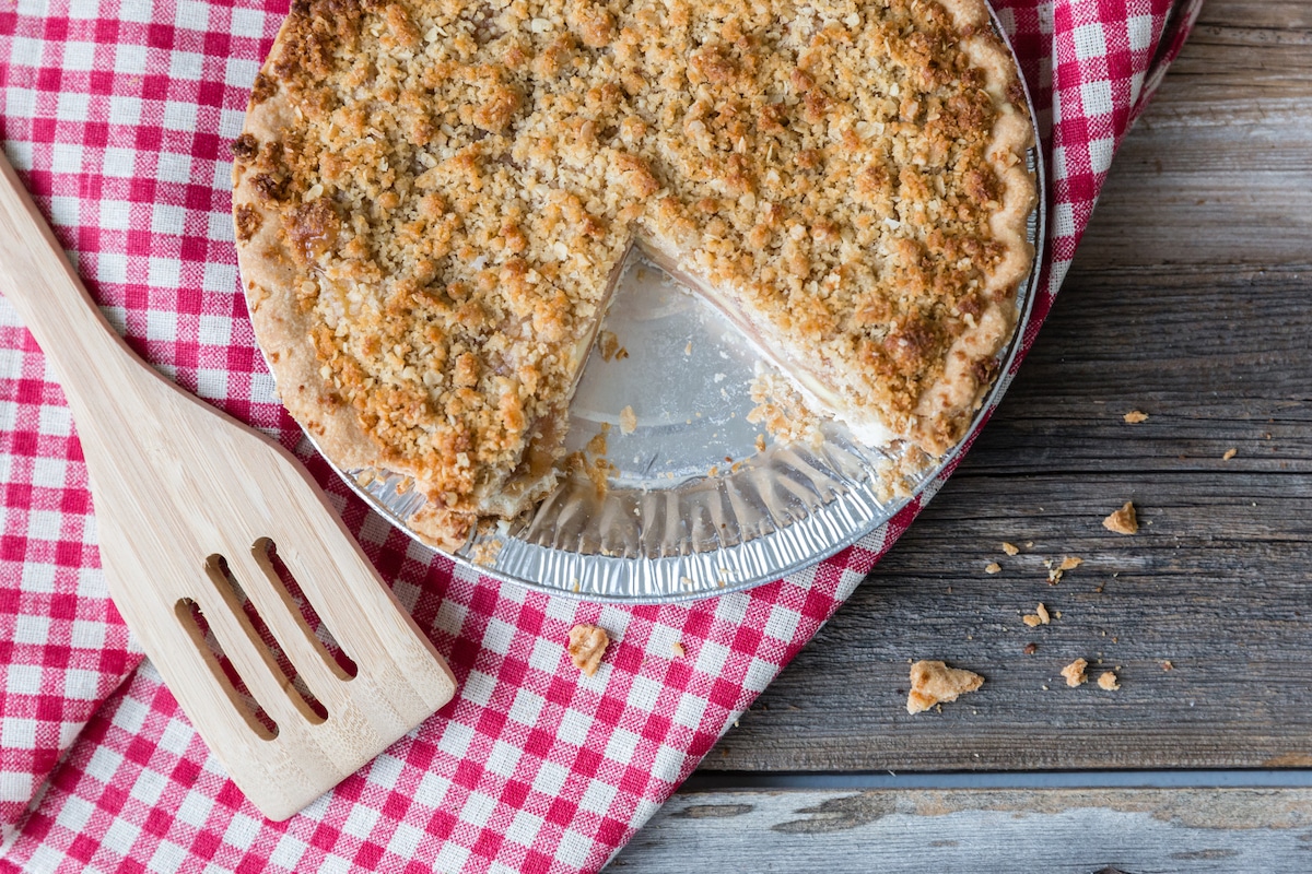 horizontal image of a whole apple crumble pie with portion removed sitting on a red checkered cloth.