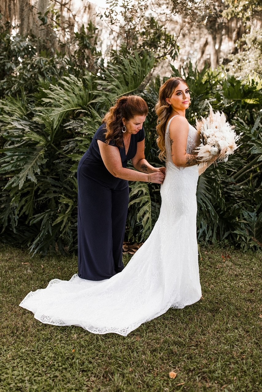 Mother of the Bride Wearing Stylish Jumpsuit While Buttoning Bride's Wedding Dress