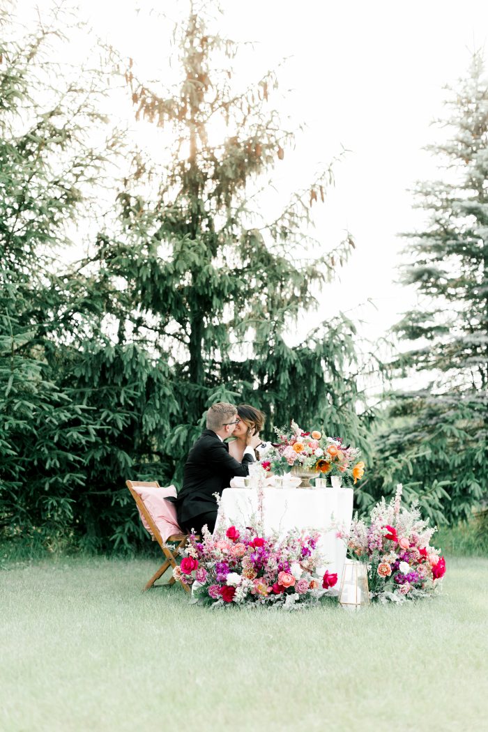 Groom Kissing Bride Wearing Maggie Sottero Wedding Dress and Sitting at Rustic Bohemian Reception Table with Vibrant Florals