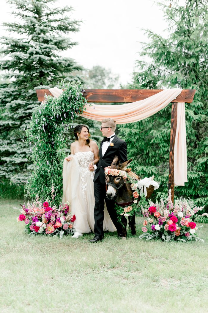 Groom Walking with Donkey and Bride Under Archway While Bride Wearing Blush Floral Wedding Dress Called Zareen by Maggie Sottero