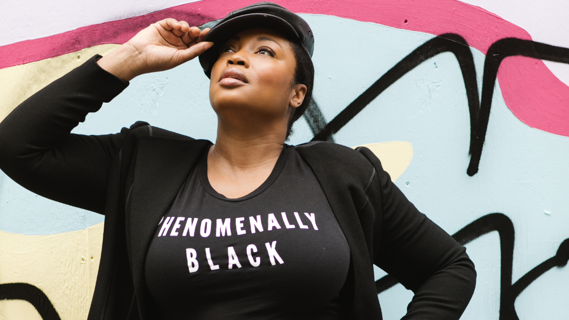 Plus Size Black Model Posing While Wearing Black T-shirt That Says "Phenomenally Black" Against Colored BackDrop